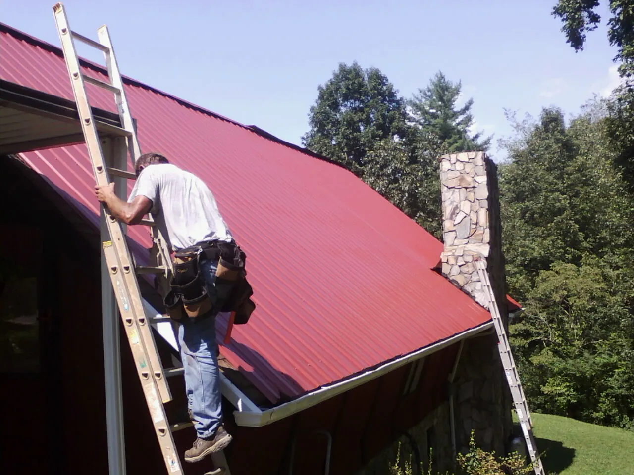 Roofer on a ladder beside a red metal roof and stone chimney — Soffit & Fascia Repair by Roofing Brothers in Marysville